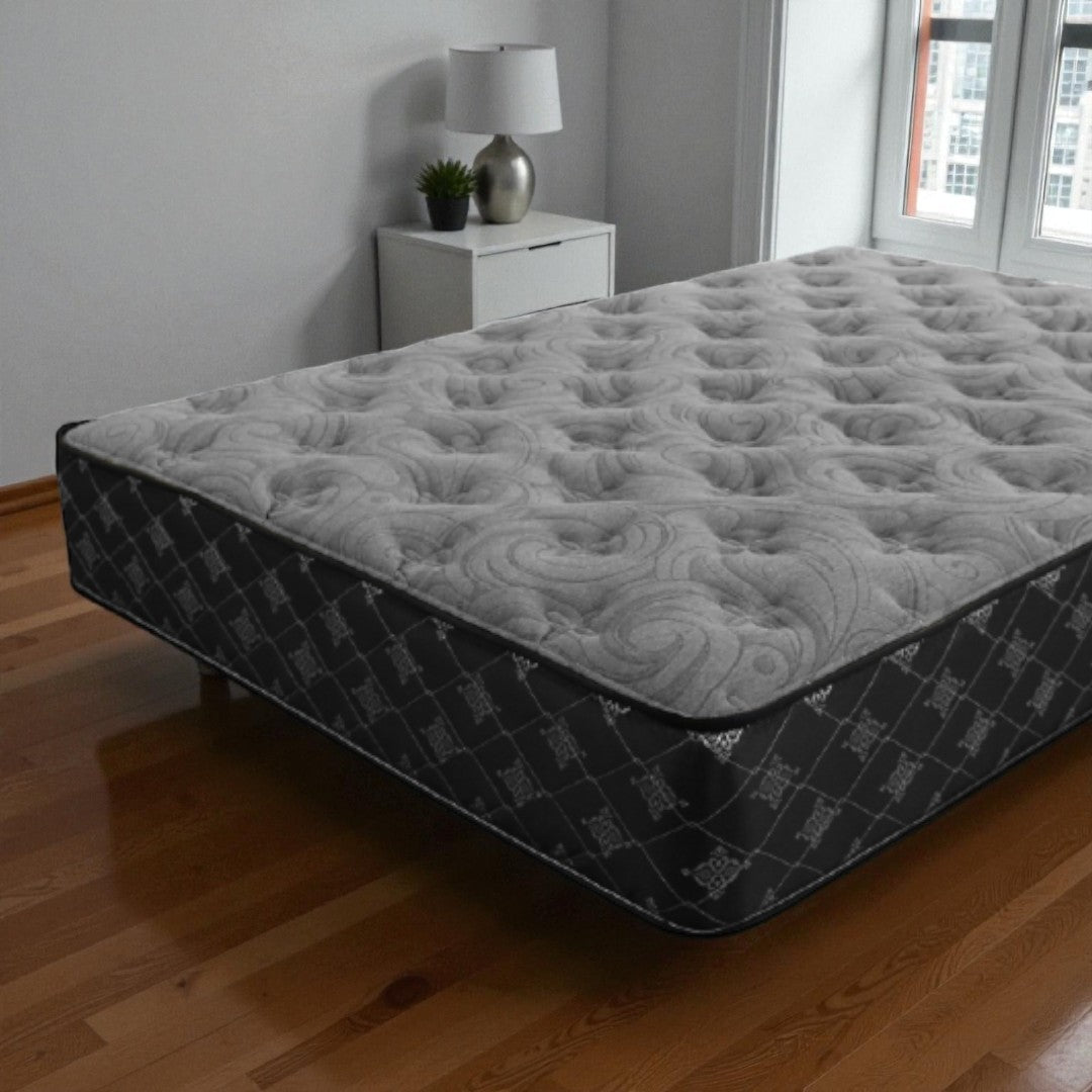 Gray mattress on a wooden floor with a nightstand and lamp in the background.
